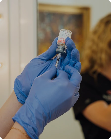 A close-up of a medical professional’s hands in blue surgical gloves carefully drawing a clear liquid into a syringe from a glass vial.
