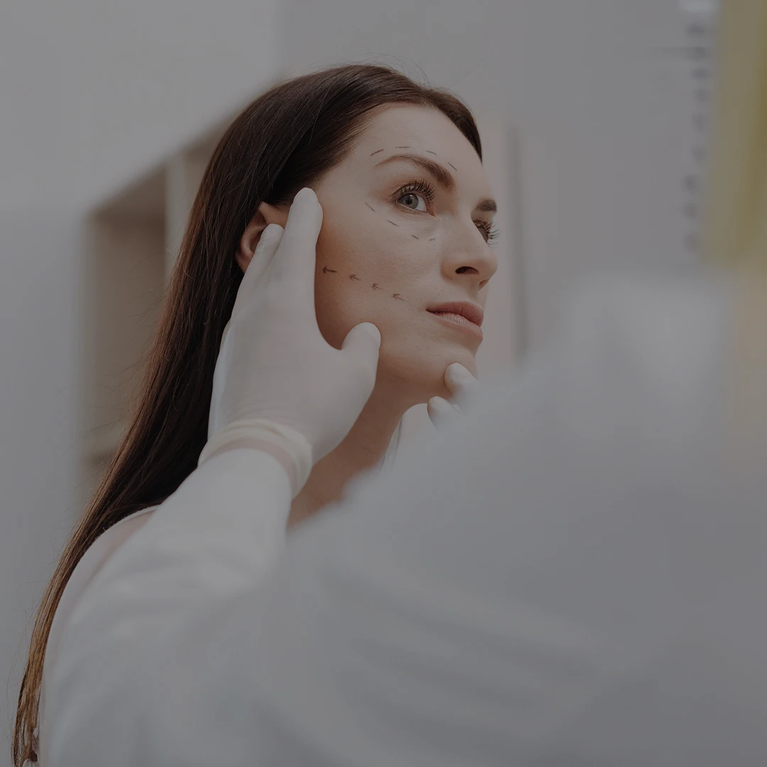 A side profile of a woman with surgical marking lines on her cheek and jawline being evaluated by a surgeon wearing white medical gloves.