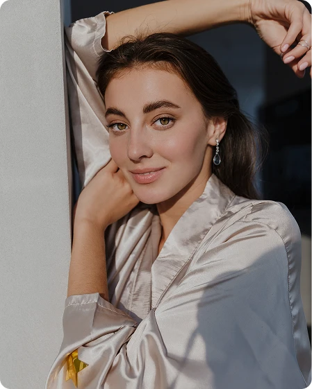 A young woman with glowing skin and brown hair wearing a silk robe poses elegantly with her hand behind her head in soft, natural light.