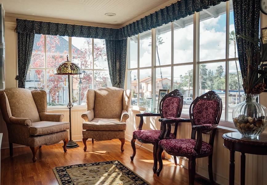 A warm, sunlit waiting room with vintage-style floral armchairs, a classic stained-glass floor lamp, and large windows overlooking a street.