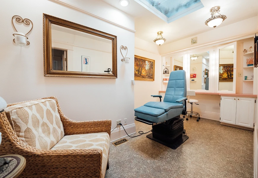 A clean medical consultation room featuring a blue adjustable procedure chair, a large ornate gold-framed mirror, and wicker seating.