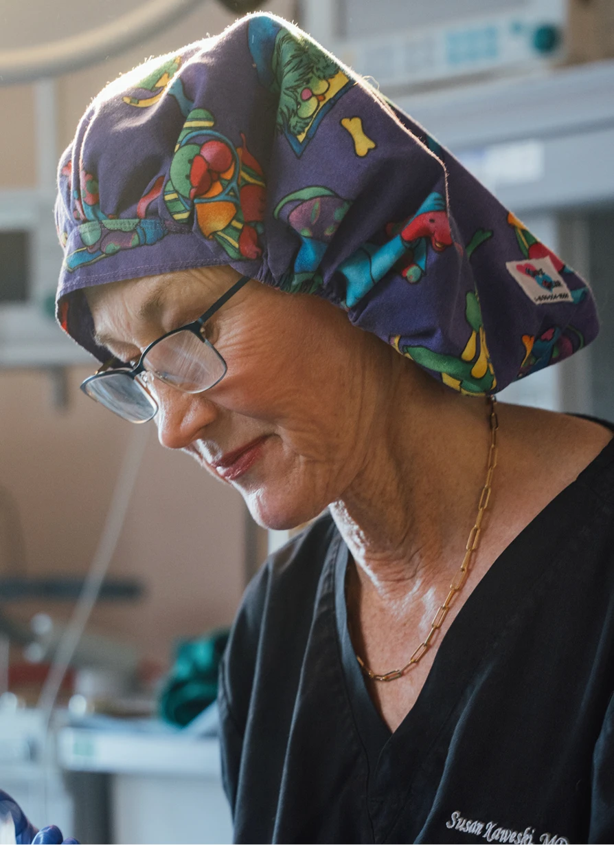 A close-up shot of Dr. Kaweski in a surgical environment, wearing a colorful patterned scrub cap and blue gloves. She is carefully examining a patient's face under bright surgical lights.