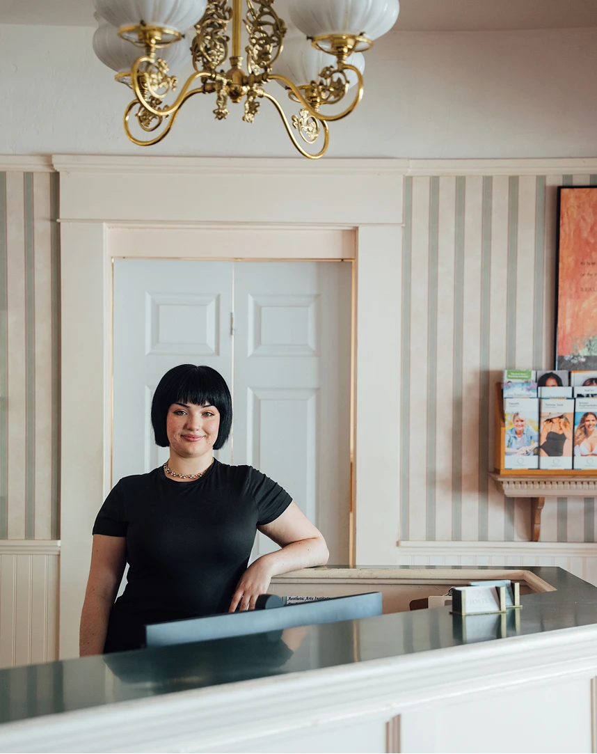 A friendly receptionist with dark hair standing behind a white marble front desk in a classically styled office with striped wallpaper and a gold chandelier.