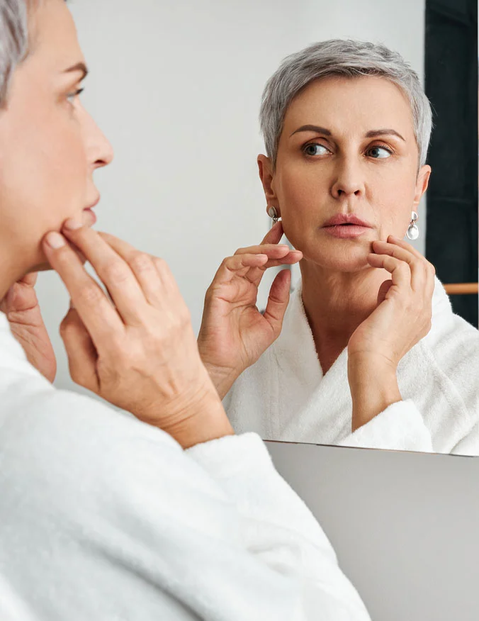 An older woman in a white robe looking in a mirror and touching her jawline, showcasing the results of skin tightening and resurfacing treatments.
