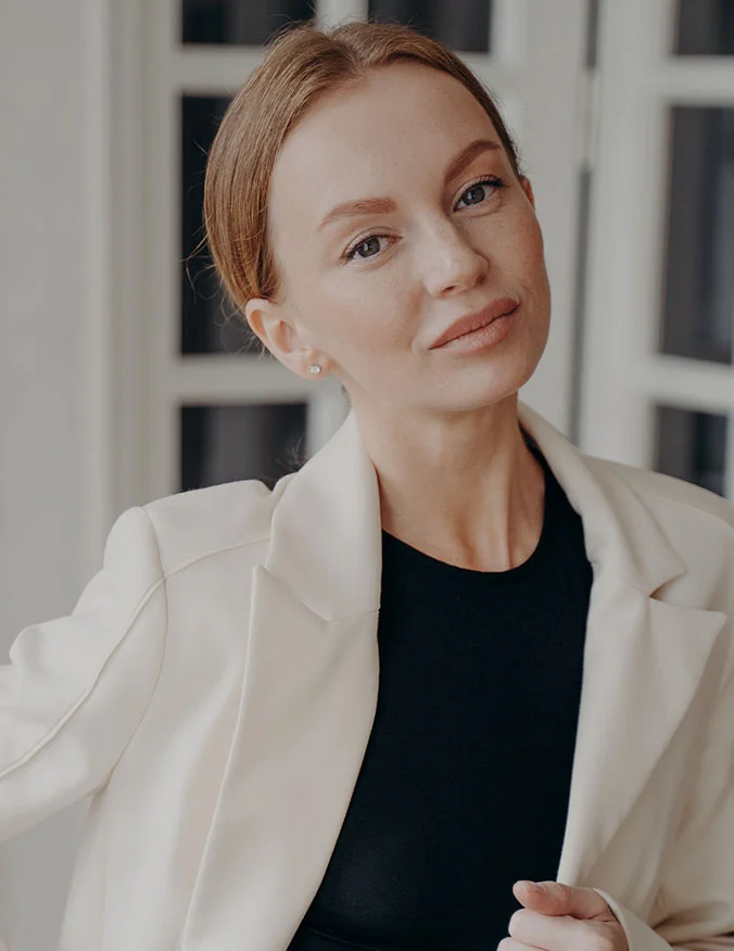 A portrait of a woman with her hair pulled back, wearing a cream blazer and black top. She has a smooth, radiant complexion, representing the results of non-surgical skin tightening.