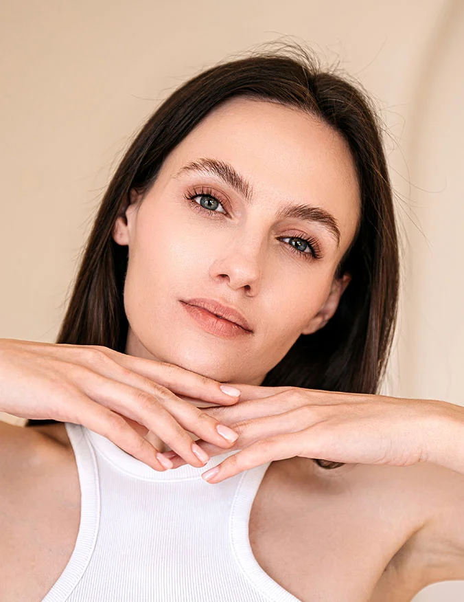 A close-up beauty shot of a woman with her hands under her chin. She has clear skin and perfectly groomed eyebrows, looking directly at the camera to showcase a youthful, refreshed appearance.