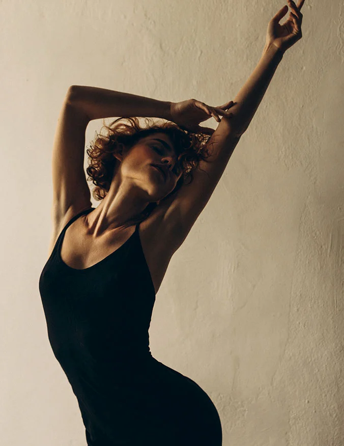 An artistic silhouette of a woman with curly hair stretching her arms upward, wearing a black bodysuit against a warm, neutral background.