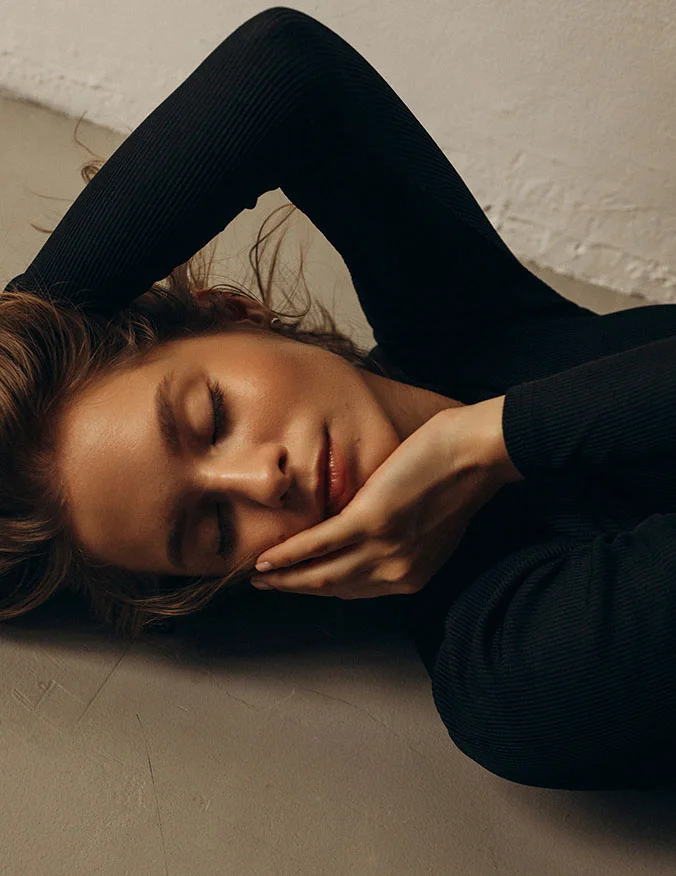A woman lying down with her eyes closed and hand to her face, wearing a black ribbed top, emphasizing defined cheekbones and facial contours.