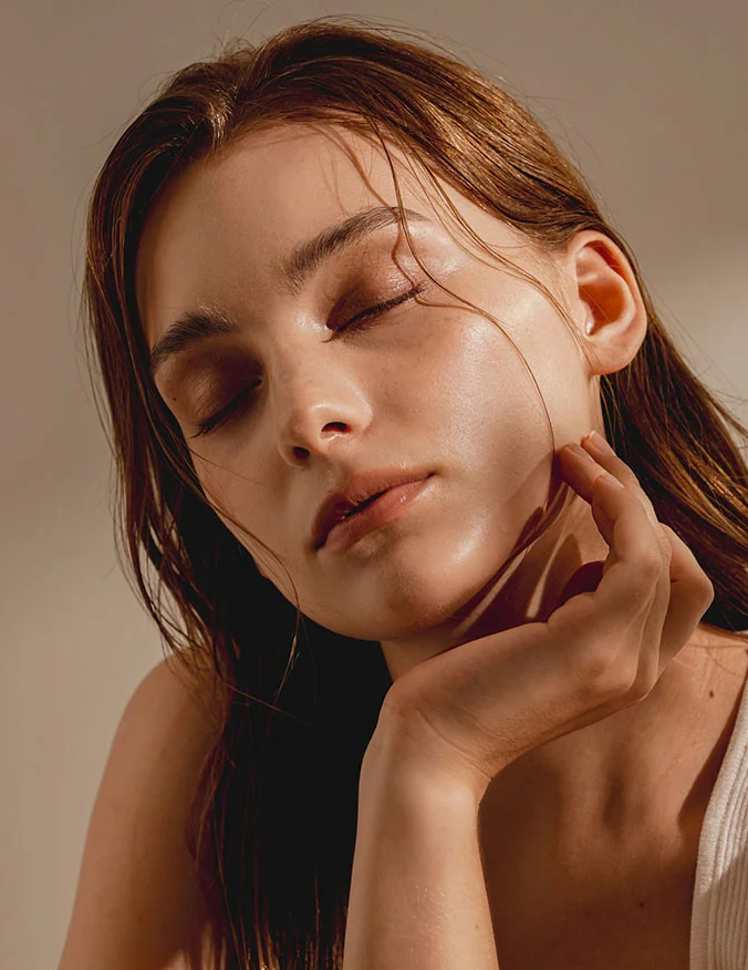 A close-up of a woman with her eyes closed and head tilted back, with wet hair and a hand near her ear, highlighting facial symmetry.