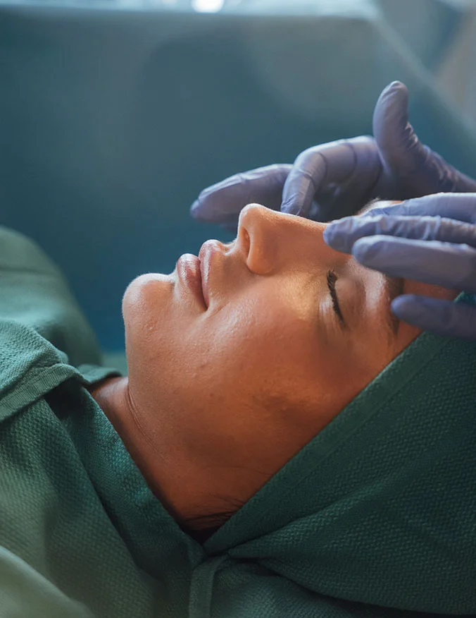 A close-up, profile view of a patient lying down in a surgical suite while a medical professional in blue gloves gently examines the eye and brow area.