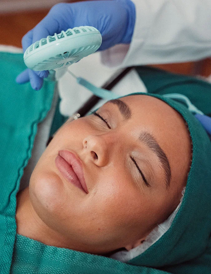 A patient with her eyes closed receiving a facial treatment, with a medical professional using a small handheld cooling fan to soothe the skin.