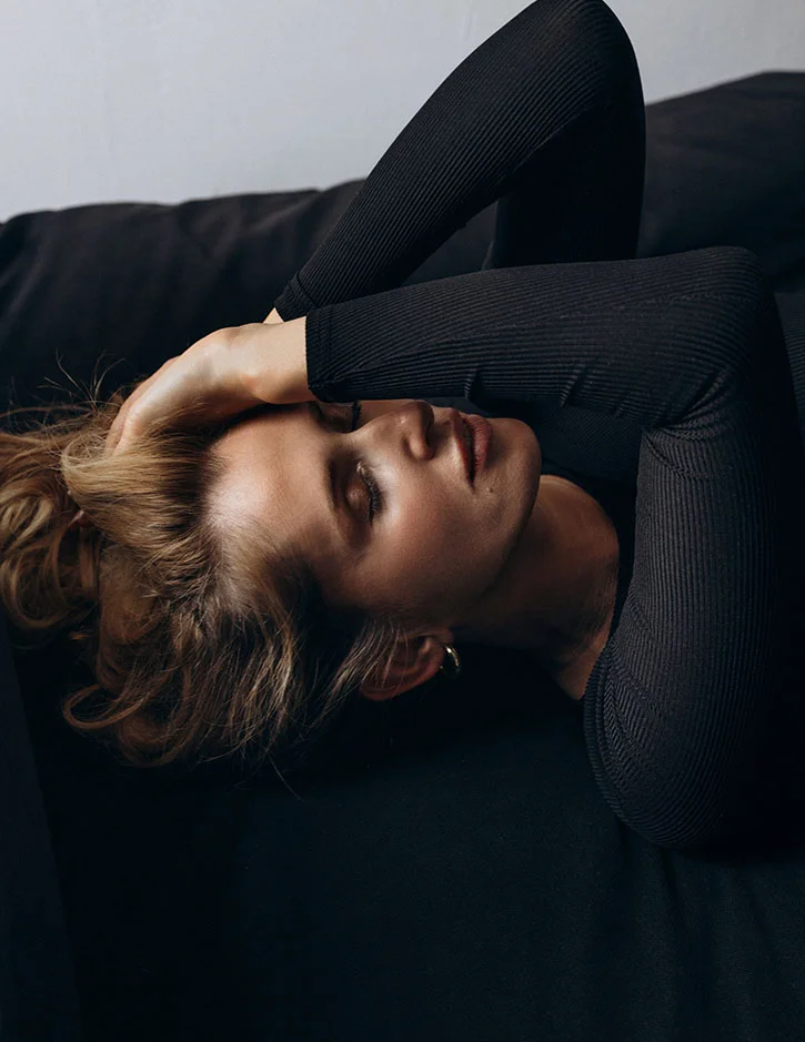 A moody, top-down shot of a woman lying back on a dark surface. She has her hands tucked under her head and is wearing a black ribbed long-sleeve top, appearing relaxed with her eyes closed.