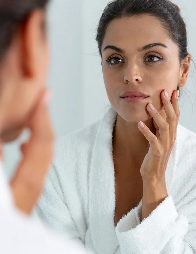 A woman in a white robe looking at her reflection in a mirror and touching her cheek, inspecting her clear and even skin tone.