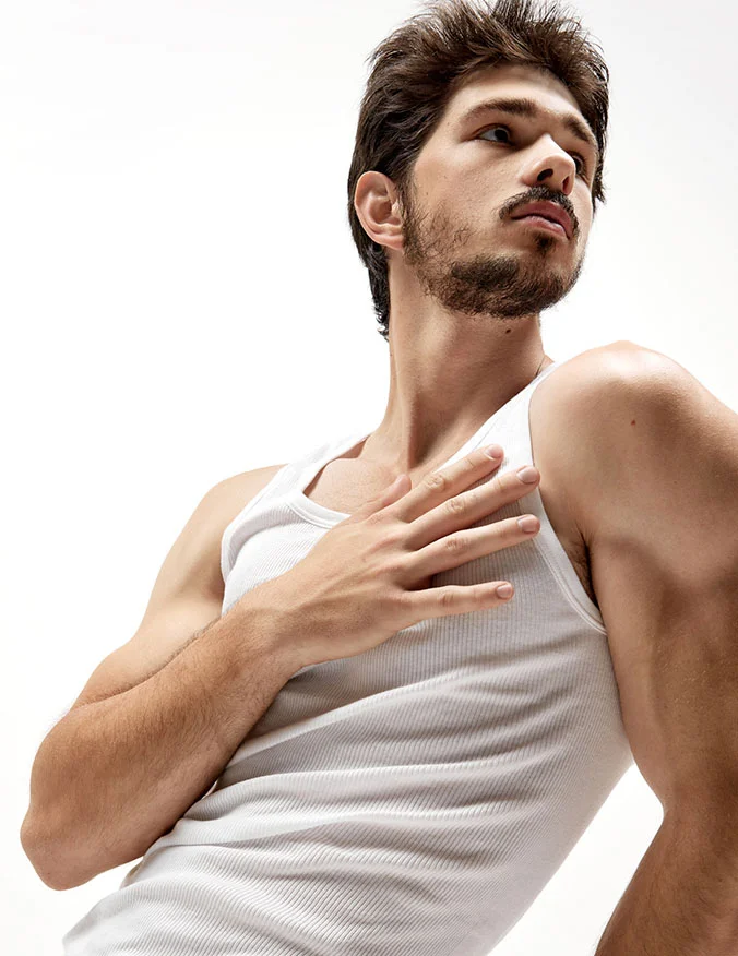 A man with a beard wearing a white ribbed tank top, looking to the side with his hand on his chest to highlight a toned and firm upper body.