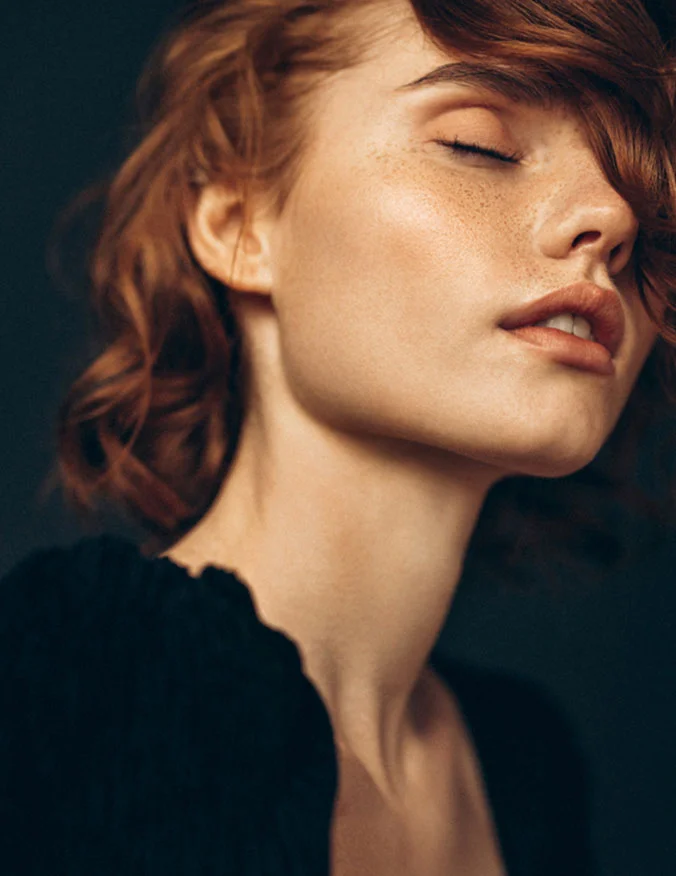 A beauty shot of a woman with red hair and freckles looking upward with her eyes closed, showcasing clear and glowing skin.