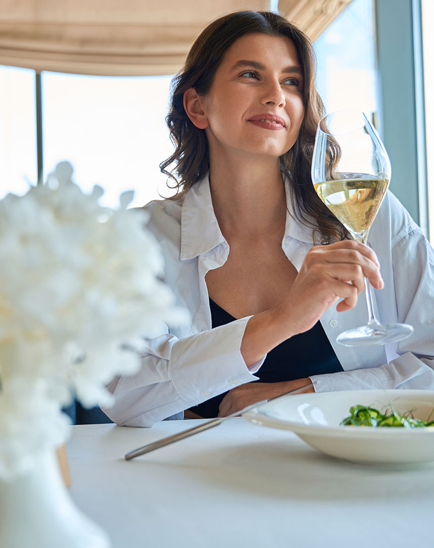 A lifestyle photo of a smiling woman sitting at a restaurant table, holding a glass of white wine. She is wearing an open white button-down shirt over a black top, with a blurred floral arrangement in the foreground.