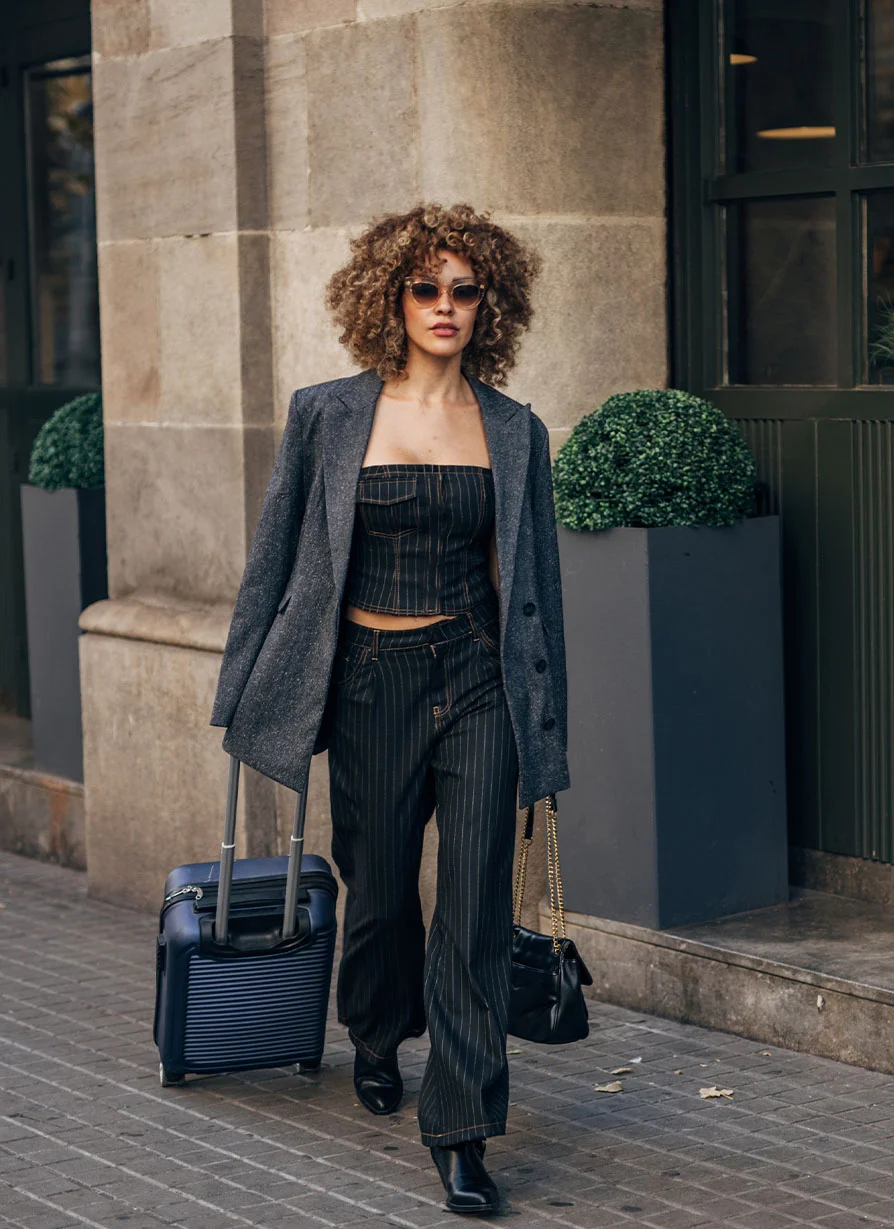 A stylish woman with curly hair walking down a city sidewalk, wearing a pinstripe suit and sunglasses while wheeling a blue carry-on suitcase, representing a traveling patient.