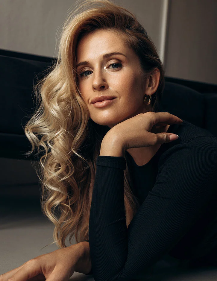 A portrait of a woman with long, wavy blonde hair resting her chin on her hand. She is wearing a black ribbed turtleneck and looking directly at the camera with a soft, confident expression.