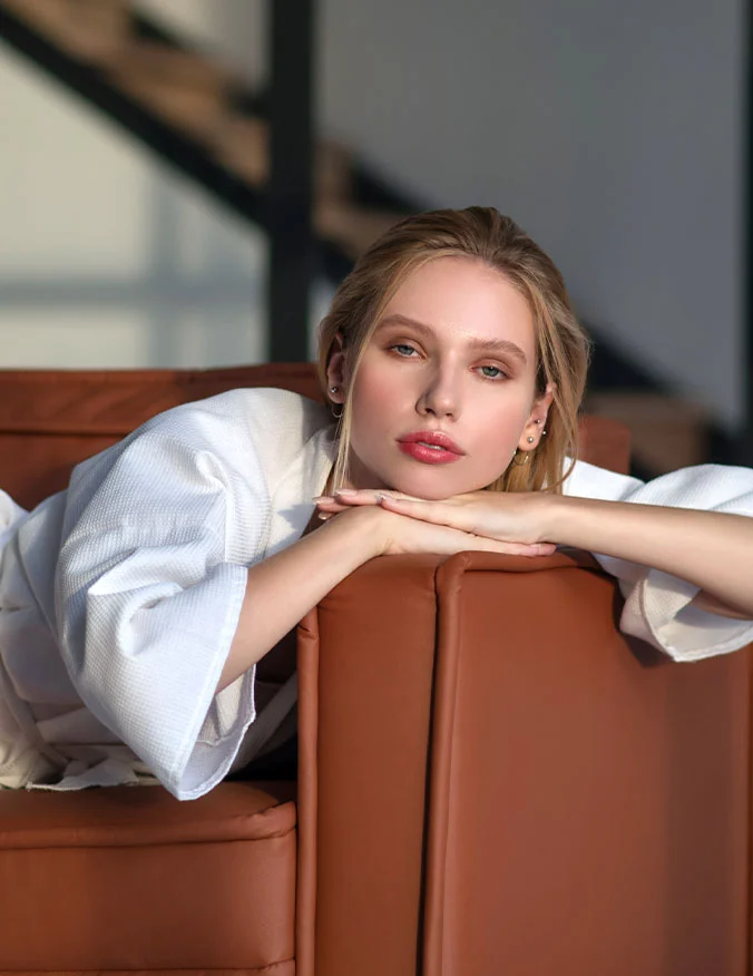 A woman in a white robe leaning over the back of a leather couch, looking directly at the camera with radiant, rejuvenated skin.