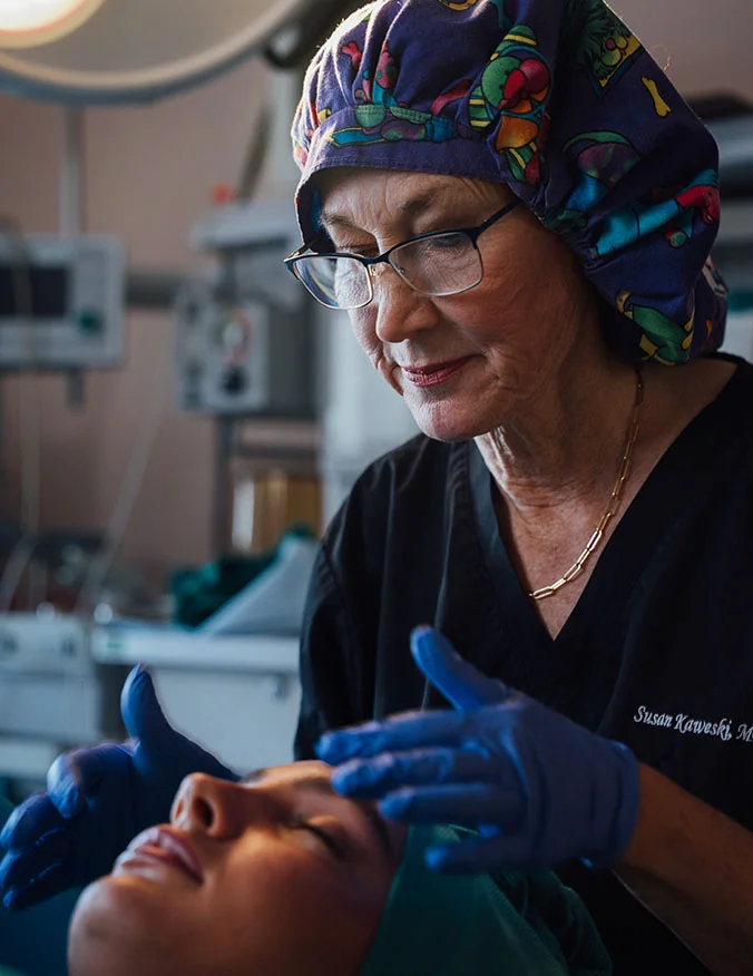A close-up shot of Dr. Kaweski in a surgical environment, wearing a colorful patterned scrub cap and blue gloves. She is carefully examining a patient's face under bright surgical lights.