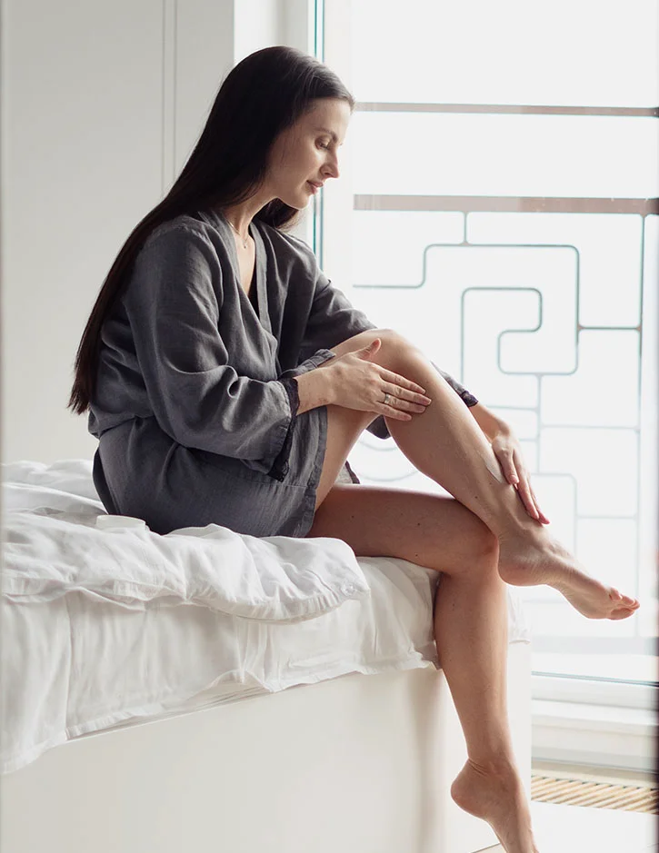 A woman in a grey linen robe sitting on the edge of a white bed by a bright window. she is gently applying cream to her legs, highlighting a self-care or post-treatment routine.