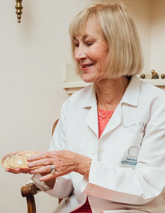 Dr. Susan Kuchli, a female plastic surgeon in a white lab coat, smiles while holding and demonstrating a clear silicone breast implant.