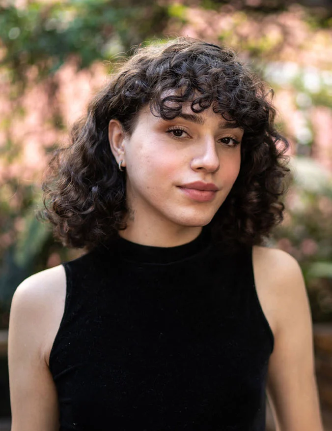 A portrait of a person with curly dark hair and a black sleeveless top, looking at the camera with a gentle expression against a blurred outdoor background.