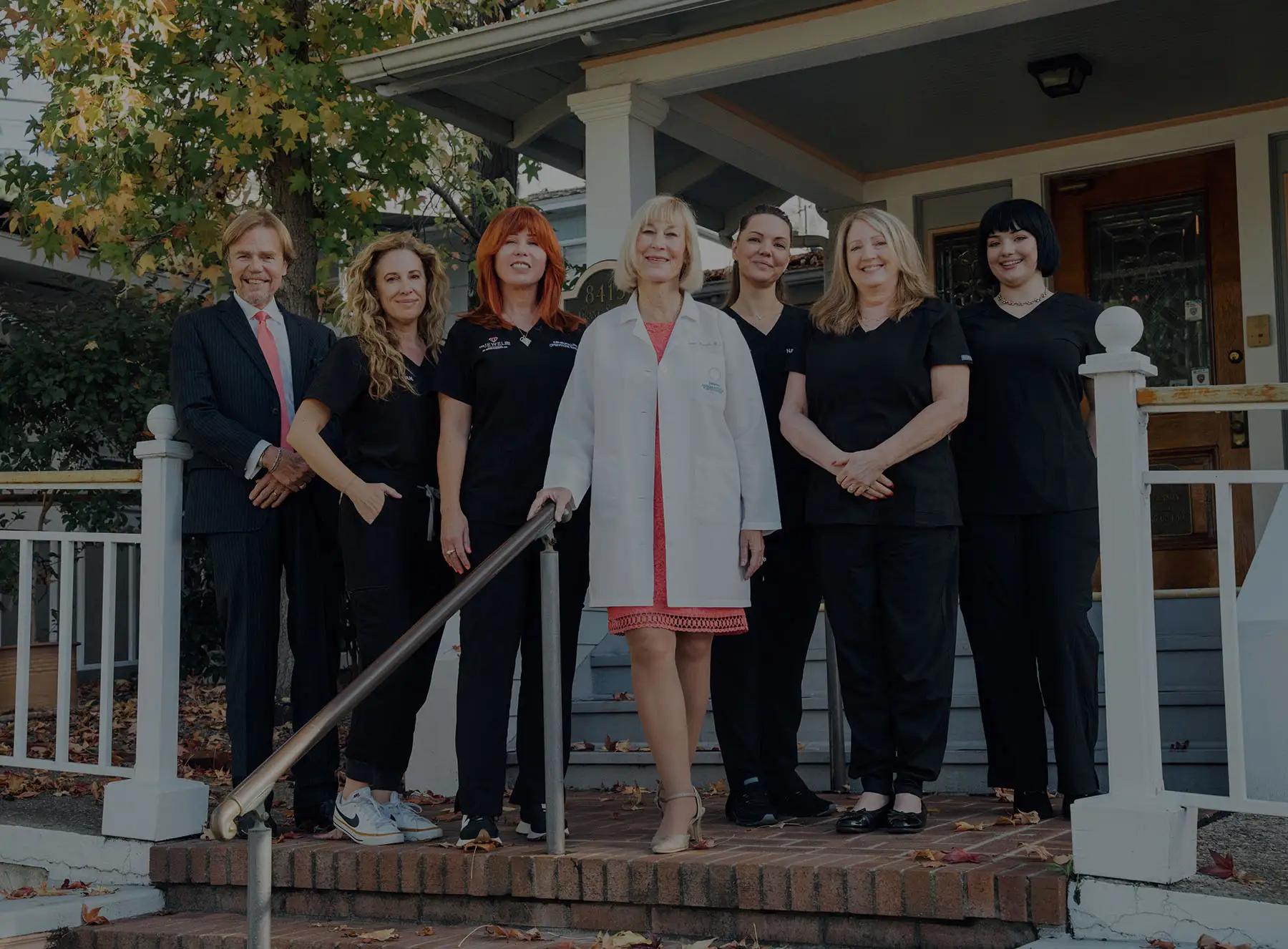 A full group portrait of Dr. Kaweski and her team standing on the brick steps of their office. Dr. Kaweski is at the center in a white coat, surrounded by her professional medical and administrative staff in black scrubs and business attire.