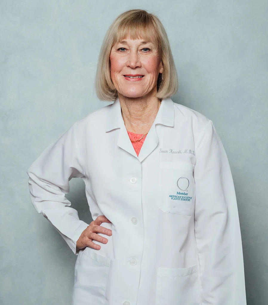 A professional portrait of Dr. Susan Kaweski against a neutral grey background. She is smiling and wearing a white lab coat with a "Member of the American Society of Plastic Surgeons" logo on the pocket.