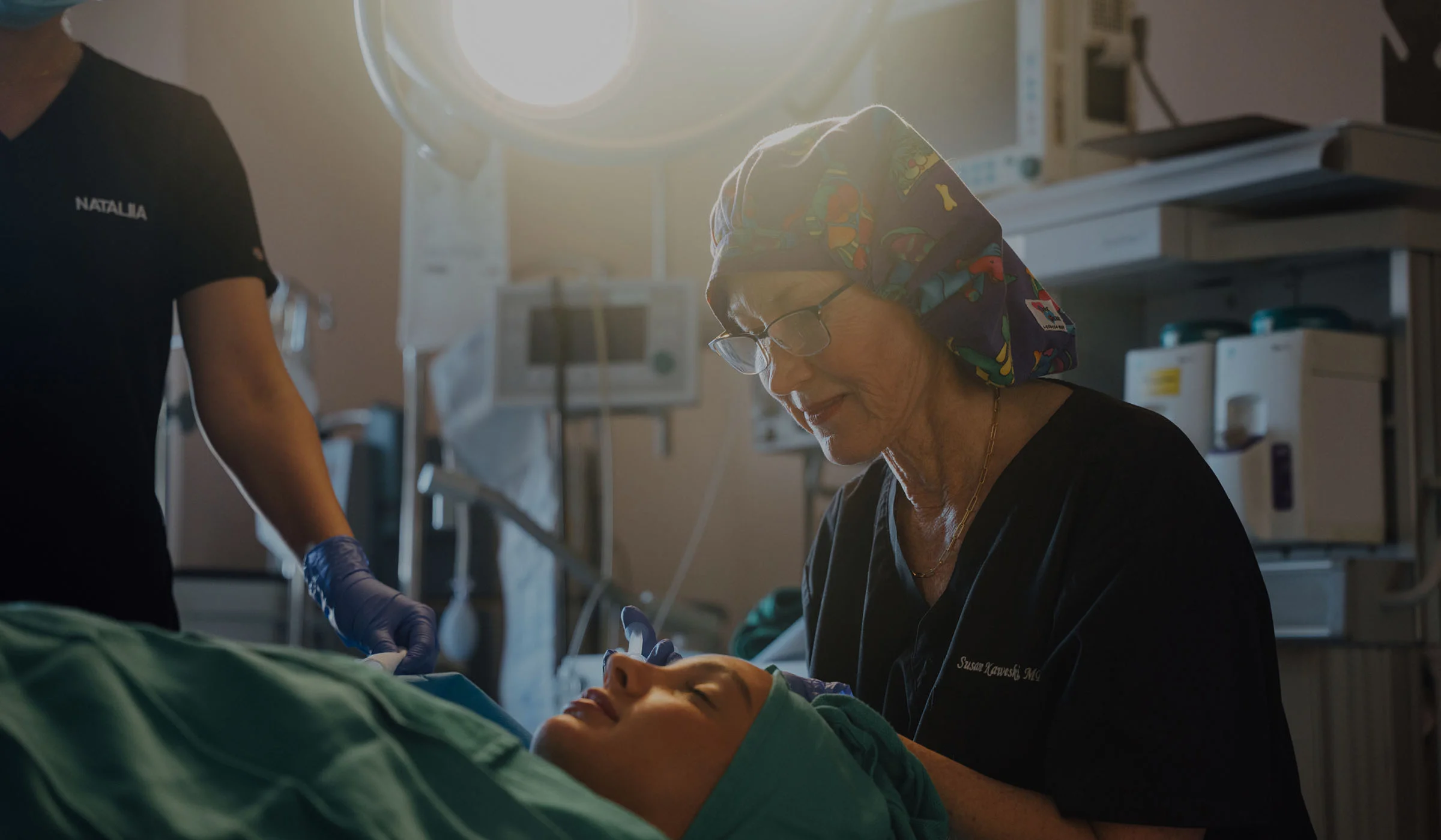 Dr. Susan Kaweski, wearing a surgical cap and glasses, focused on a patient during a fat grafting procedure in an operating room.