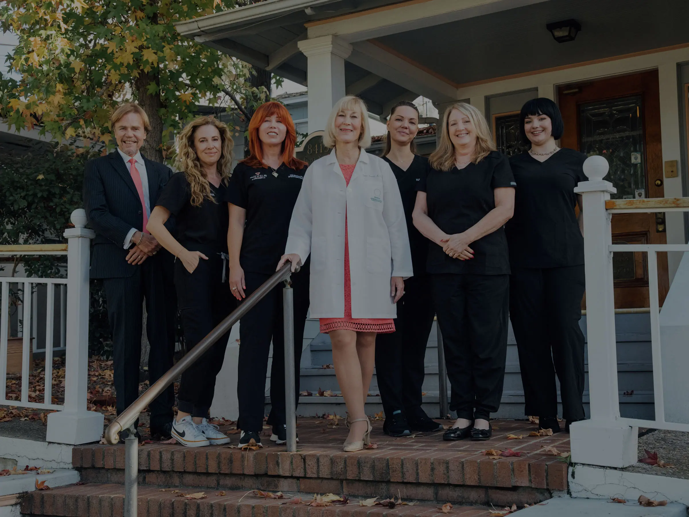 A full group portrait of Dr. Kaweski and her team standing on the brick steps of their office. Dr. Kaweski is at the center in a white coat, surrounded by her professional medical and administrative staff in black scrubs and business attire.
