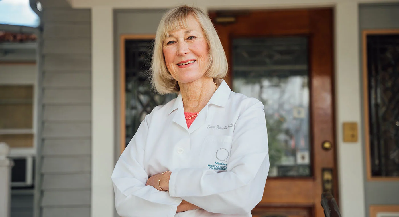 Dr. Susan Kaweski posing confidently with her arms crossed in front of a classic wooden door and grey siding, representing the professional yet welcoming exterior of her practice.