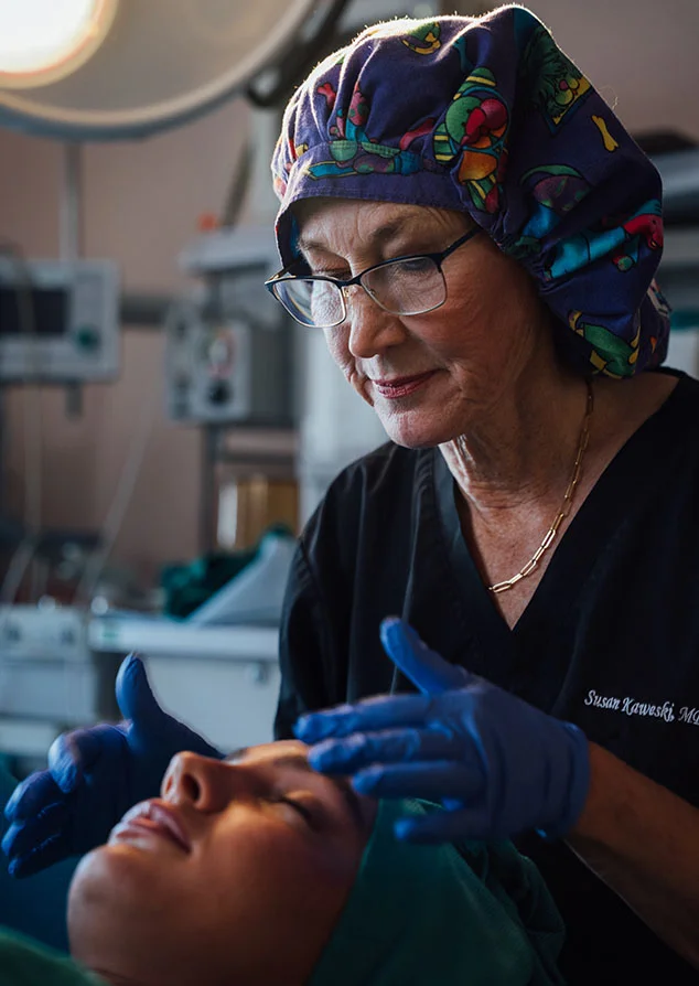 A close-up shot of Dr. Kaweski in a surgical environment, wearing a colorful patterned scrub cap and blue gloves. She is carefully examining a patient's face under bright surgical lights.